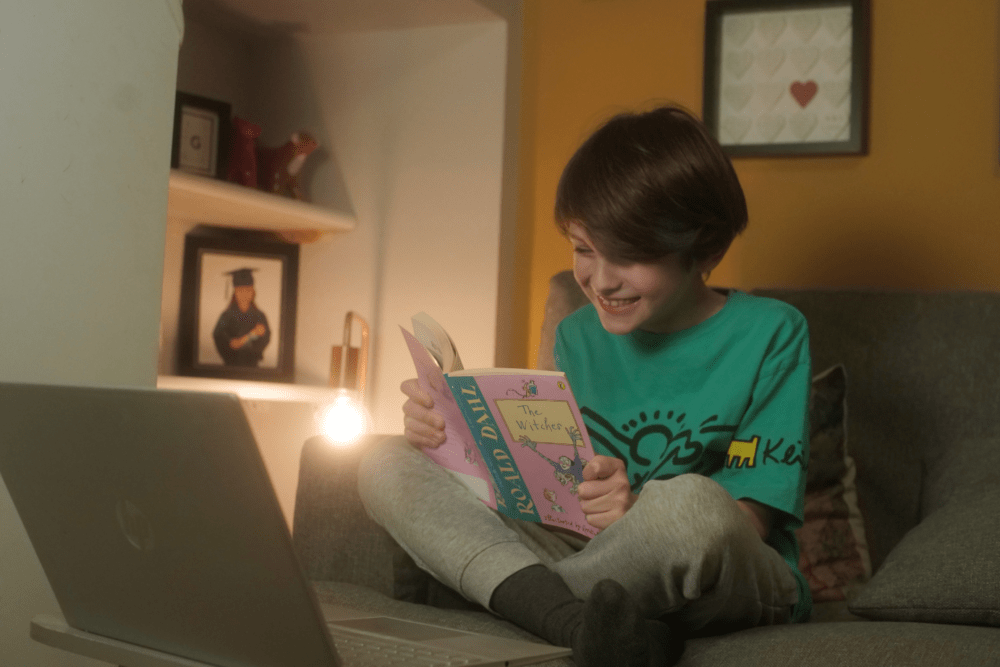 Child reads book in front of laptop where his volunteer reads with him